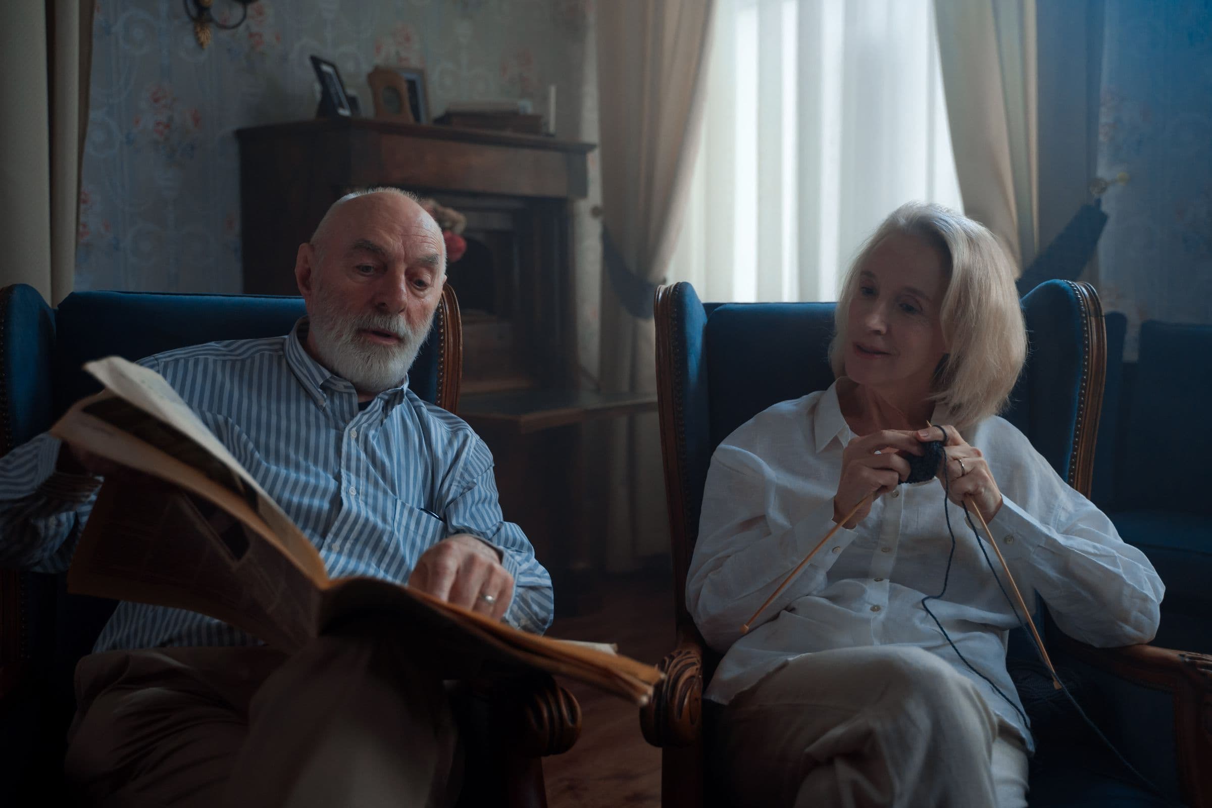 Older couple relaxing in armchairs in the club lounge, reading and knitting